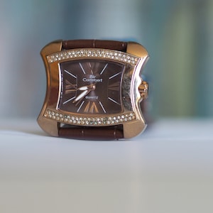 May include: A brown leather wristwatch with a gold-tone case and a diamond-encrusted bezel. The watch face is brown with gold-tone hands and Roman numeral hour markers. The watch face also has the text "Rolex Cotebert QUARTZ".