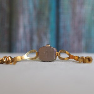 May include: Gold-tone wristwatch with a square face and a metal band. The back of the watch is visible, showing the watch's mechanism.