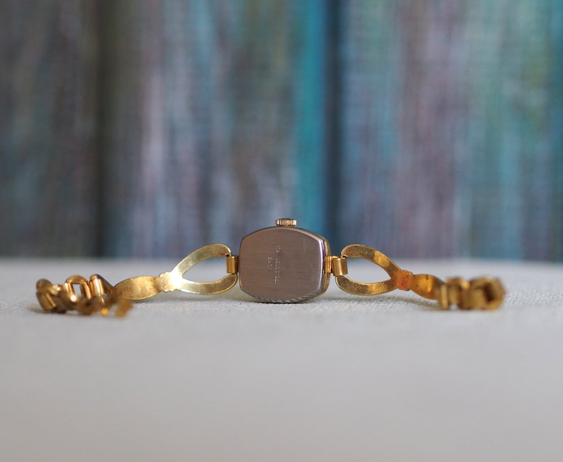 May include: Gold-tone wristwatch with a square face and a metal band. The back of the watch is visible, showing the watch's mechanism.