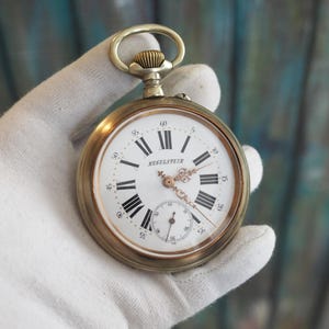 May include: A silver pocket watch with Roman numeral markings on the face. The watch face is white with black markings and the words "Regulateur" printed in black. The watch has a gold-toned case.