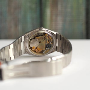 May include: A silver wristwatch with a stainless steel band. The back of the watch is open, revealing the inner workings of the watch.