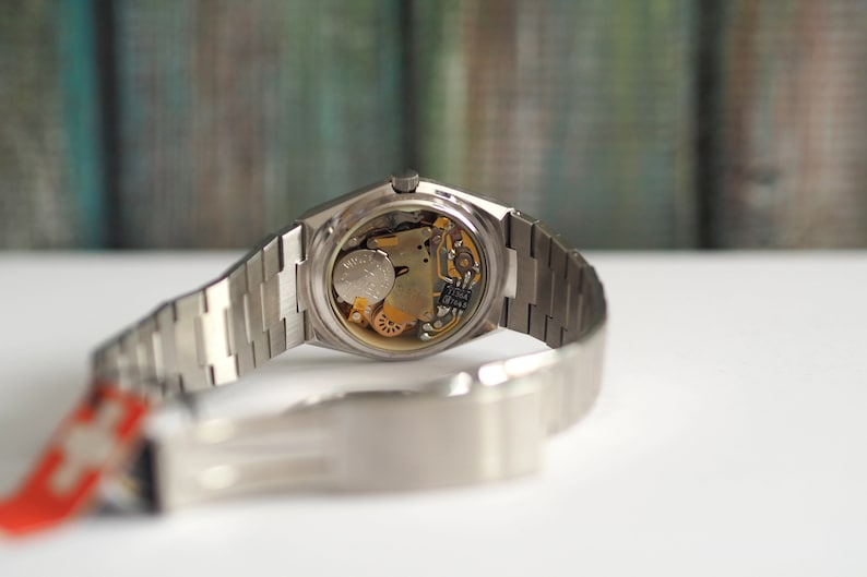 May include: A silver wristwatch with a stainless steel band. The back of the watch is open, revealing the inner workings of the watch.