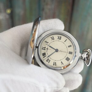 Puede incluir: Un reloj de bolsillo plateado con números romanos en la esfera. La esfera del reloj es blanca con agujas negras y la palabra "ultrama" impresa en negro.