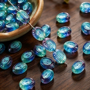 May include: Oval glass beads featuring a translucent blue and purple gradient with a pressed sunburst or floral pattern on the surface. Some beads are strung on a thin wire, while others are scattered loosely around a ceramic bowl.