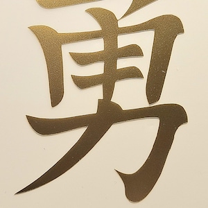 Op de afbeelding: Een goudkleurig Japans Kanji-symbool voor "Moed" op een witte achtergrond. Het woord "Courage" staat in een lichtblauw, cursief lettertype onder het symbool. Het symbool is een gestileerd, kalligrafisch ontwerp.