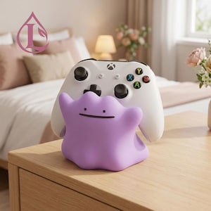 Puede incluir: Un soporte para mando de color morado claro con forma de mancha amorfa, que presenta un rostro sencillo con dos pequeños puntos como ojos y una línea horizontal como boca, sosteniendo un mando de juego inalámbrico blanco con joysticks negros.