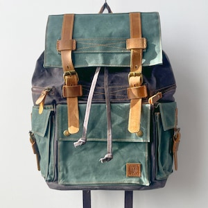 Peut inclure: Un sac à dos vert et marine avec des sangles et boucles en cuir marron. Le sac à dos comporte plusieurs poches et une fermeture à cordon. Le sac est suspendu à une chaîne sur fond blanc.