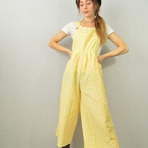 May include: A yellow and white gingham jumpsuit with wide legs. The jumpsuit has adjustable straps and a button closure. The person is wearing a black beret, a white t-shirt, and black boots. The outfit is a vintage style.