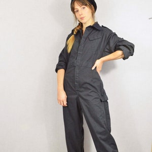 May include: A dark grey jumpsuit with long sleeves, a collared neckline, and multiple pockets. The jumpsuit has a belt at the waist and is worn with white trainers and a black beret. The overall style is casual and utilitarian.