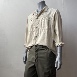 Può includere: Una camicia beige a maniche lunghe con due tasche sul petto e bottoni scuri, abbinata a pantaloni cargo verde oliva. Le maniche della camicia sono arrotolate. L'outfit è esposto su un manichino.
