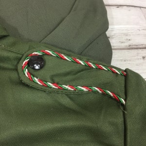 May include: Close-up of a green fabric with a red, white, and green braided trim. A black button with a star design is sewn onto the fabric.