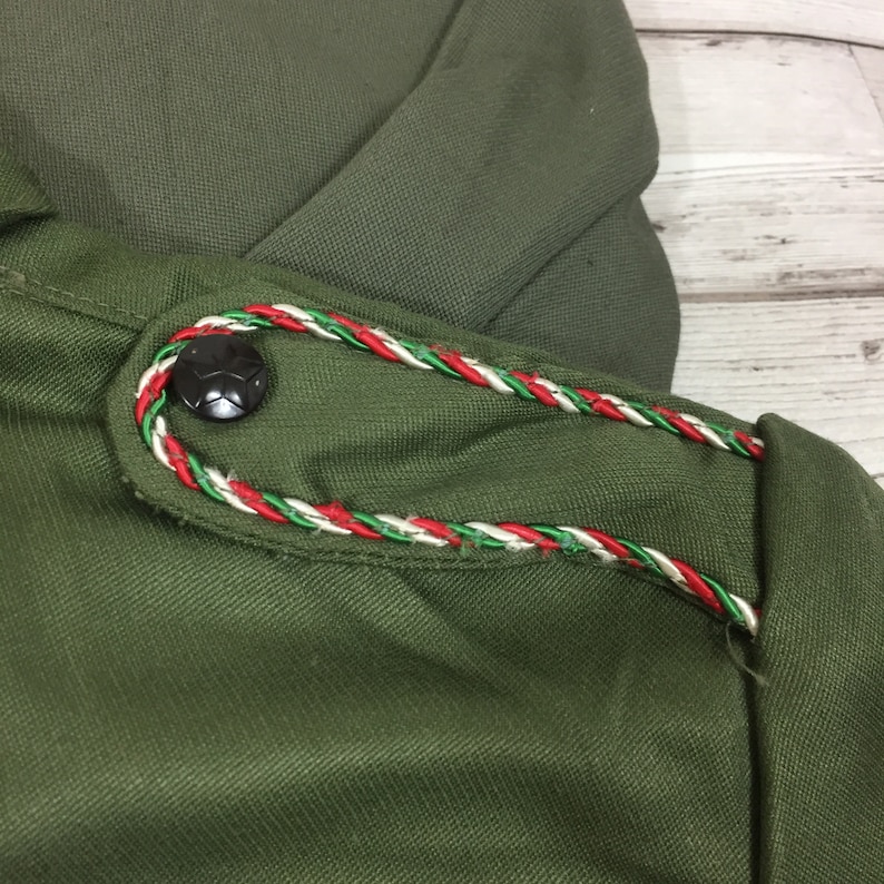 May include: Close-up of a green fabric with a red, white, and green braided trim. A black button with a star design is sewn onto the fabric.