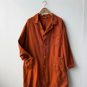 May include: A long-sleeved, burnt orange coat featuring a collared neckline, a button-down front closure, one chest pocket, and two lower patch pockets. The garment is constructed from a textured fabric and hangs from a wooden hanger.
