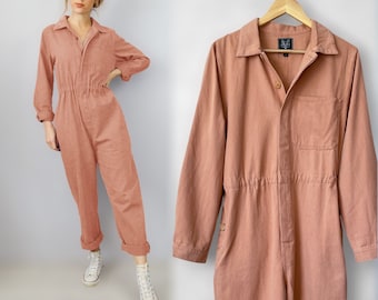 Puede incluir: Un mono de color rosa polvoriento con mangas largas, cuello de camisa y cierre de botón. El mono tiene la cintura ajustada y un bolsillo de parche en el lado izquierdo.