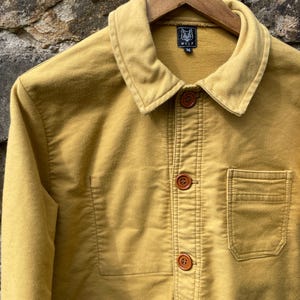 May include: Mustard-colored jacket with a collared neckline and wooden buttons. The jacket features a chest pocket and the brand logo "WOLF" on a tag. The jacket is hanging on a wooden hanger.
