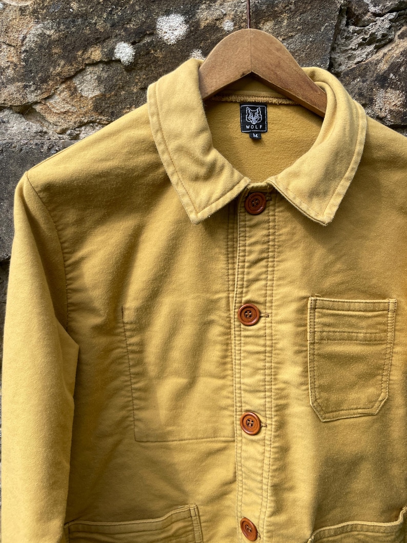 May include: Mustard-colored jacket with a collared neckline and wooden buttons. The jacket features a chest pocket and the brand logo "WOLF" on a tag. The jacket is hanging on a wooden hanger.