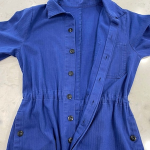 May include: A cobalt blue jumpsuit with a collared neckline and long sleeves. The jumpsuit features a button-down front, a drawstring waist, and pockets. The fabric has a subtle vertical texture. Black buttons and a single pocket add detail to the garment.