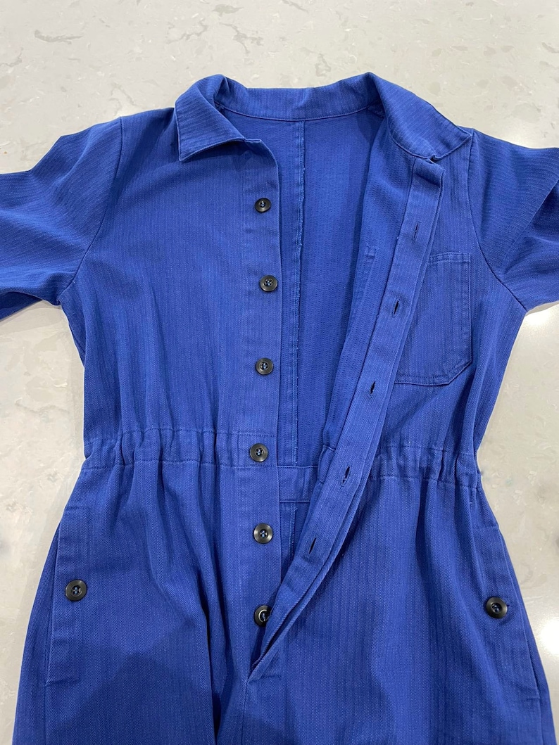 May include: A cobalt blue jumpsuit with a collared neckline and long sleeves. The jumpsuit features a button-down front, a drawstring waist, and pockets. The fabric has a subtle vertical texture. Black buttons and a single pocket add detail to the garment.