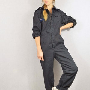 May include: Dark grey jumpsuit with a button-up front, a belt, and a small patch on the chest. The jumpsuit is paired with a black beret and white high-top trainers. The outfit is a vintage style.