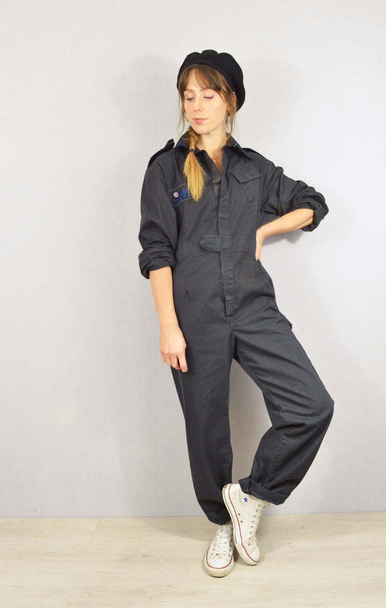 May include: Dark grey jumpsuit with a button-up front, a belt, and a small patch on the chest. The jumpsuit is paired with a black beret and white high-top trainers. The outfit is a vintage style.