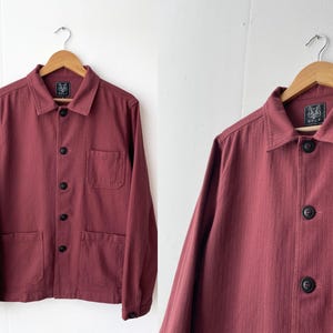 Op de afbeelding: Een bordeauxrood werkjasje met een knoopsluiting, twee borstzakken en een enkele zak op de linkermouw. Het jasje heeft een geweven label met het woord "WOLF" aan de linkerkant van de kraag.