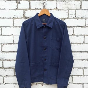 Op de afbeelding: Een marineblauw jack met lange mouwen, een klassieke kraag, een knoopsluiting met donkere knopen, een borstzak en twee opgestikte zakken aan de onderzijde. Het jack hangt aan een houten kledinghanger en heeft een label in de nek.