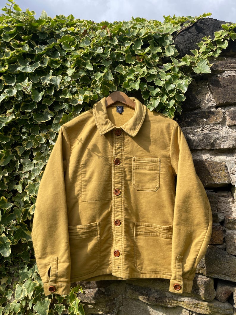 May include: Mustard-colored jacket with a button-up front, two chest pockets, and two lower pockets. The jacket has a collared neckline and buttoned cuffs. The garment is hanging on a wooden hanger against a stone wall and ivy.