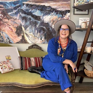 May include: A woman wearing a blue jumpsuit, a gray hat, and glasses sits on a green sofa. A large landscape painting hangs on the wall behind her. A colorful rug and a wooden shelf with pottery are also visible.