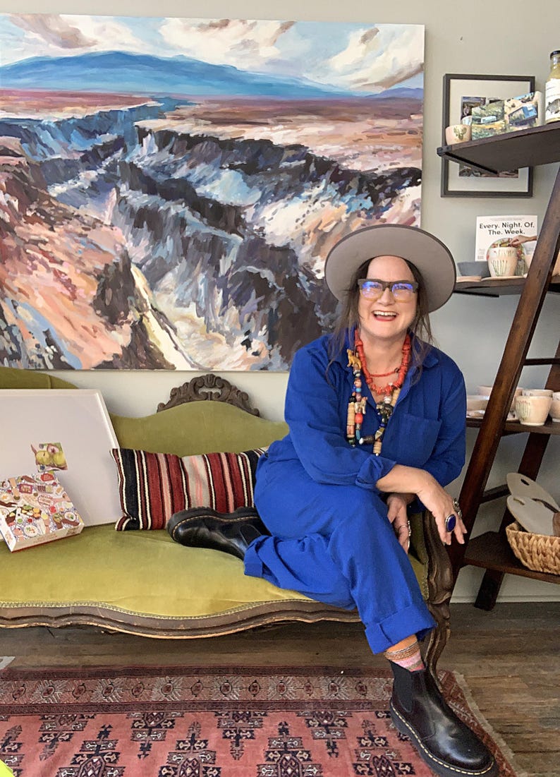May include: A woman wearing a blue jumpsuit, a gray hat, and glasses sits on a green sofa. A large landscape painting hangs on the wall behind her. A colorful rug and a wooden shelf with pottery are also visible.