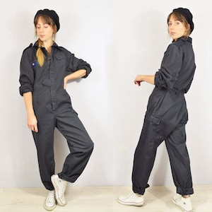 Pode incluir: Macacão cinza escuro com mangas compridas, gola e vários bolsos. O macacão tem uma frente abotoada e é combinado com uma boina preta e tênis brancos. O estilo geral é casual e utilitário.