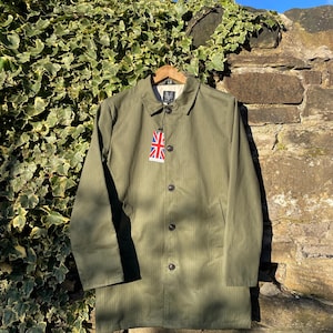 Op de afbeelding: Een olijfgroene jas met visgraatpatroon, voorzien van een klassieke kraag, lange mouwen en een knoopsluiting aan de voorzijde. Een klein label met de Union Jack vlag hangt aan de bovenste knoop en een merklabel is zichtbaar in de kraag.