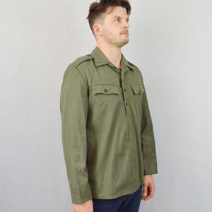 May include: A long-sleeved, olive green shirt with a button-up front and a single pocket on the left chest. The shirt has a collar with a pointed tip and a button closure.