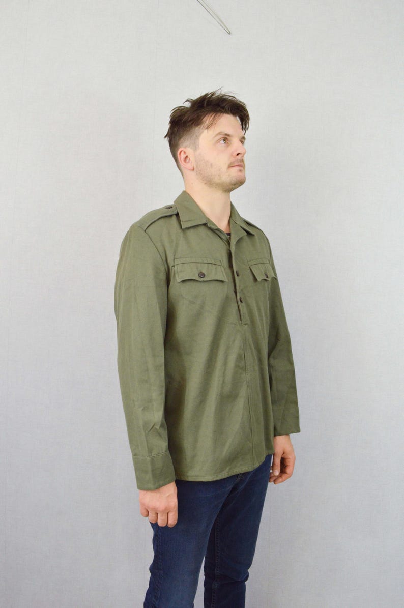 May include: A long-sleeved, olive green shirt with a button-up front and a single pocket on the left chest. The shirt has a collar with a pointed tip and a button closure.