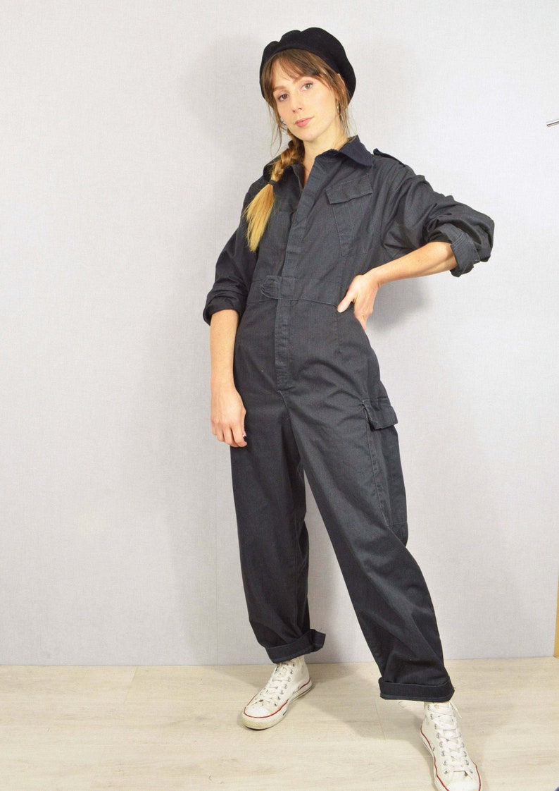 May include: A dark grey jumpsuit with long sleeves, a collared neckline, and multiple pockets. The jumpsuit has a belt at the waist and is worn with white trainers and a black beret. The overall style is casual and utilitarian.
