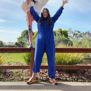 May include: A woman in a vibrant blue jumpsuit stands with arms raised, smiling. She wears colorful sneakers. Another person is partially visible, wearing a cream-colored top and pink pants, standing on a wooden fence. The background features greenery and a blue sky.