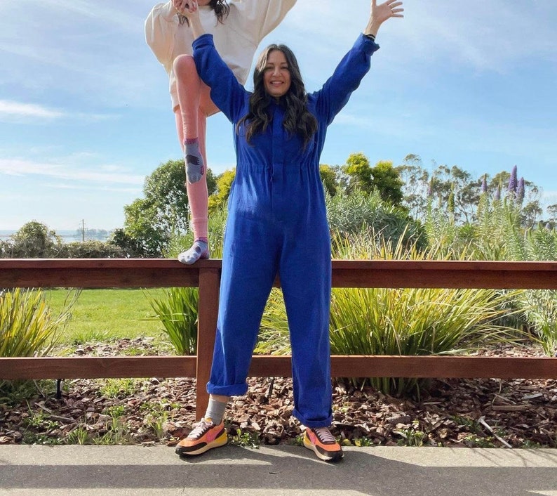 May include: A woman in a vibrant blue jumpsuit stands with arms raised, smiling. She wears colorful sneakers. Another person is partially visible, wearing a cream-colored top and pink pants, standing on a wooden fence. The background features greenery and a blue sky.