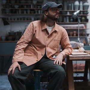 May include: A person wearing a peach-colored jacket, a gray baseball cap, and dark green pants. The jacket has a button closure and a chest pocket. The person is sitting on a stool in a workshop setting with shelves of pottery in the background.