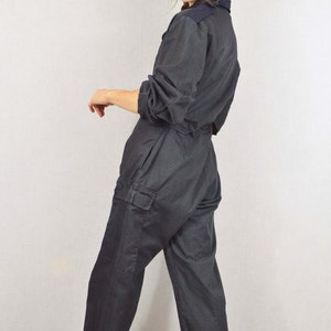 May include: Dark grey jumpsuit with a collared neckline, long sleeves, and cargo pockets. The jumpsuit is worn with a black beret and white high-top trainers. A casual, yet stylish, one-piece garment.