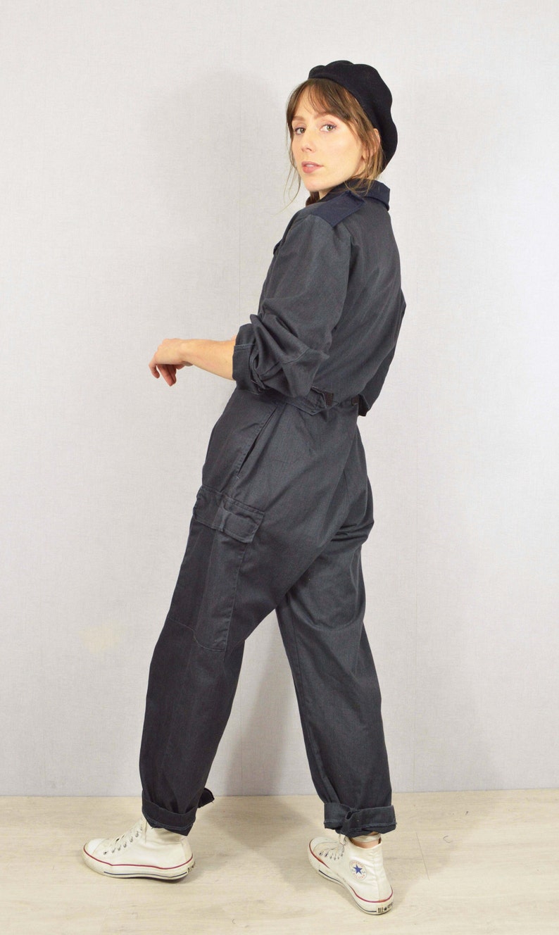 May include: Dark grey jumpsuit with a collared neckline, long sleeves, and cargo pockets. The jumpsuit is worn with a black beret and white high-top trainers. A casual, yet stylish, one-piece garment.