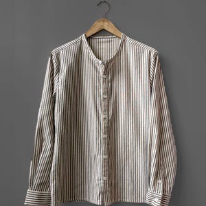 Puede incluir: Una camisa de manga larga con botones y un patrón de rayas verticales en tonos marrón, blanco y gris oscuro. La camisa presenta un cuello mao, una tapeta frontal con botones claros y puños abotonados en las mangas.