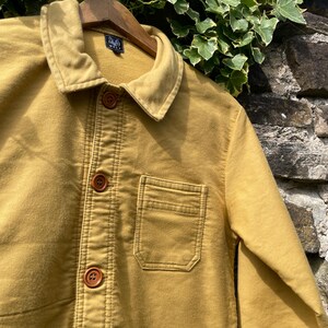 May include: Mustard-colored jacket with a button-up front, a collar, and a chest pocket. The jacket is hanging on a wooden hanger. The jacket has brown buttons and visible stitching details. The jacket is made of a soft, durable fabric.