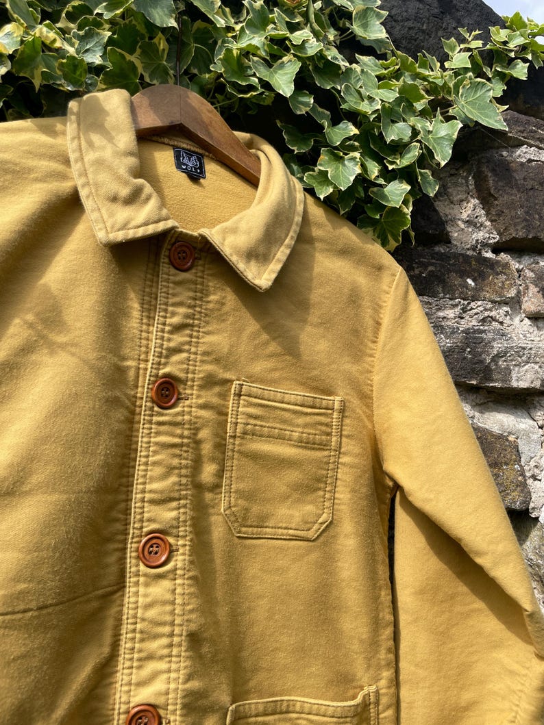 May include: Mustard-colored jacket with a button-up front, a collar, and a chest pocket. The jacket is hanging on a wooden hanger. The jacket has brown buttons and visible stitching details. The jacket is made of a soft, durable fabric.