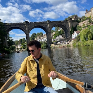 May include: A person rowing a wooden boat on a river under a stone arch bridge. The person is wearing a yellow jacket, blue jeans, and sunglasses. The sky is blue with white clouds.