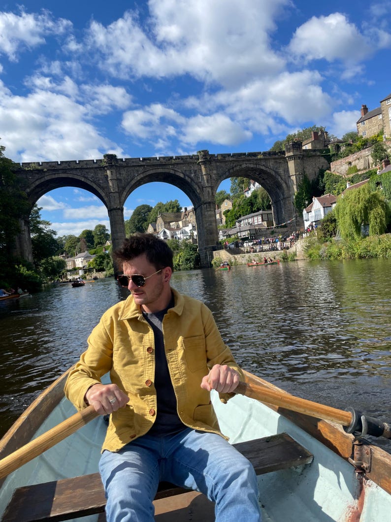May include: A person rowing a wooden boat on a river under a stone arch bridge. The person is wearing a yellow jacket, blue jeans, and sunglasses. The sky is blue with white clouds.