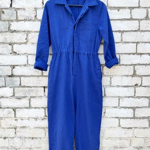 May include: A vibrant blue jumpsuit with long sleeves, a collared neckline, and a button-up front. The waist is cinched with an elastic band. The cuffs are rolled up. The jumpsuit hangs on a wooden hanger against a white brick wall.