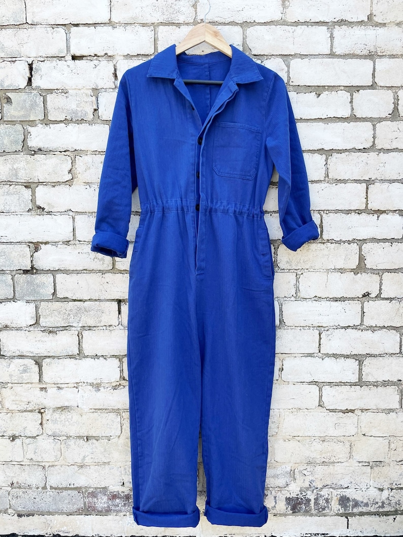 May include: A vibrant blue jumpsuit with long sleeves, a collared neckline, and a button-up front. The waist is cinched with an elastic band. The cuffs are rolled up. The jumpsuit hangs on a wooden hanger against a white brick wall.