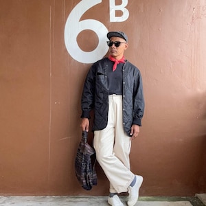 Puede incluir: Una persona con una chaqueta acolchada negra, pantalones blancos y zapatillas blancas. También lleva una gorra gris, gafas de sol y un pañuelo rojo. Sostiene un bolso tote estampado. El fondo es una pared marrón con el número "6B" en blanco.