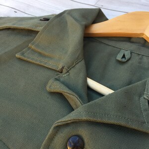 May include: Close-up of a green cotton shirt collar with a button and a metal hook. The shirt is folded and draped over a wooden hanger.