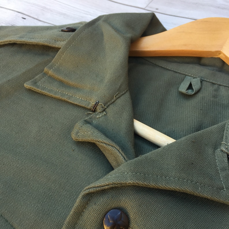 May include: Close-up of a green cotton shirt collar with a button and a metal hook. The shirt is folded and draped over a wooden hanger.