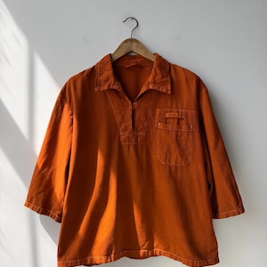 May include: A burnt orange pullover shirt with three-quarter length sleeves, a collared neckline, and a partial front button placket. The shirt features a single chest pocket with a flap closure and is displayed on a wooden hanger.